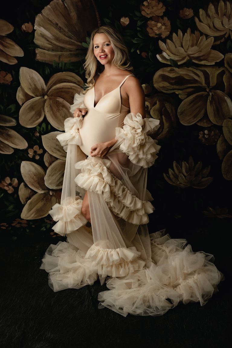Expectant mother looking incredibly glamorous in tulle cream cape in studio with dramatic floral background