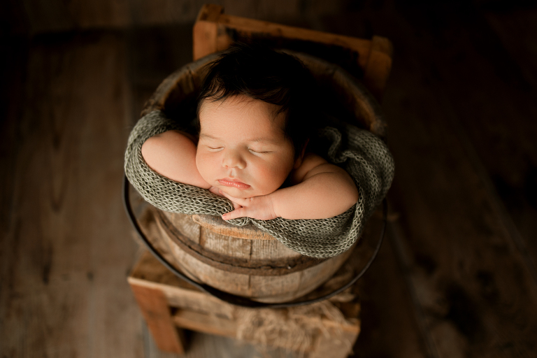 Newborn baby boy in wooden bucket on top of a wooden chair with green knit wrap to cover him