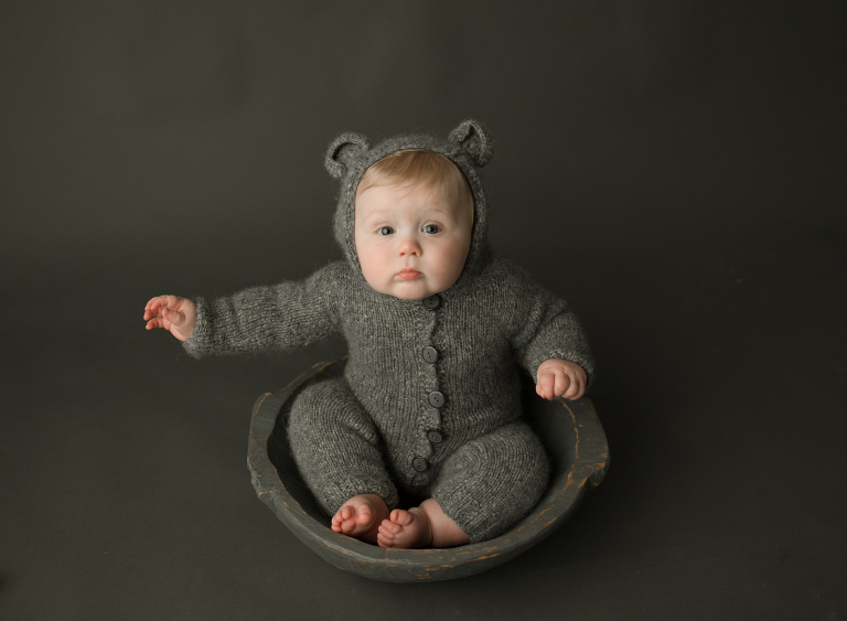 8 month old baby boy in bear onesie sitting in bowl on gray backdrop in studio