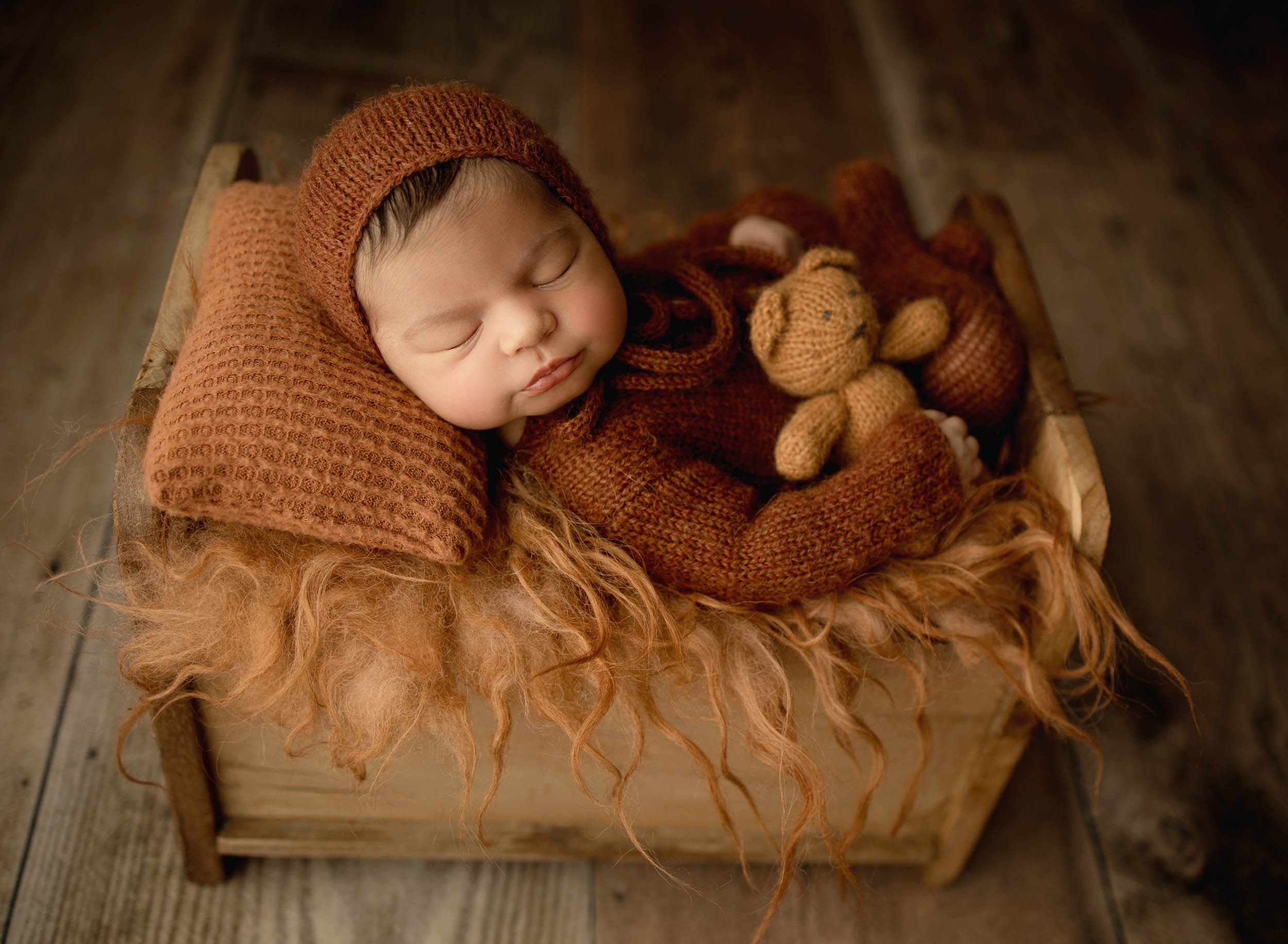 Newborn baby boy in orange knited onesie holding matching teddy bear lying in a wooden crate