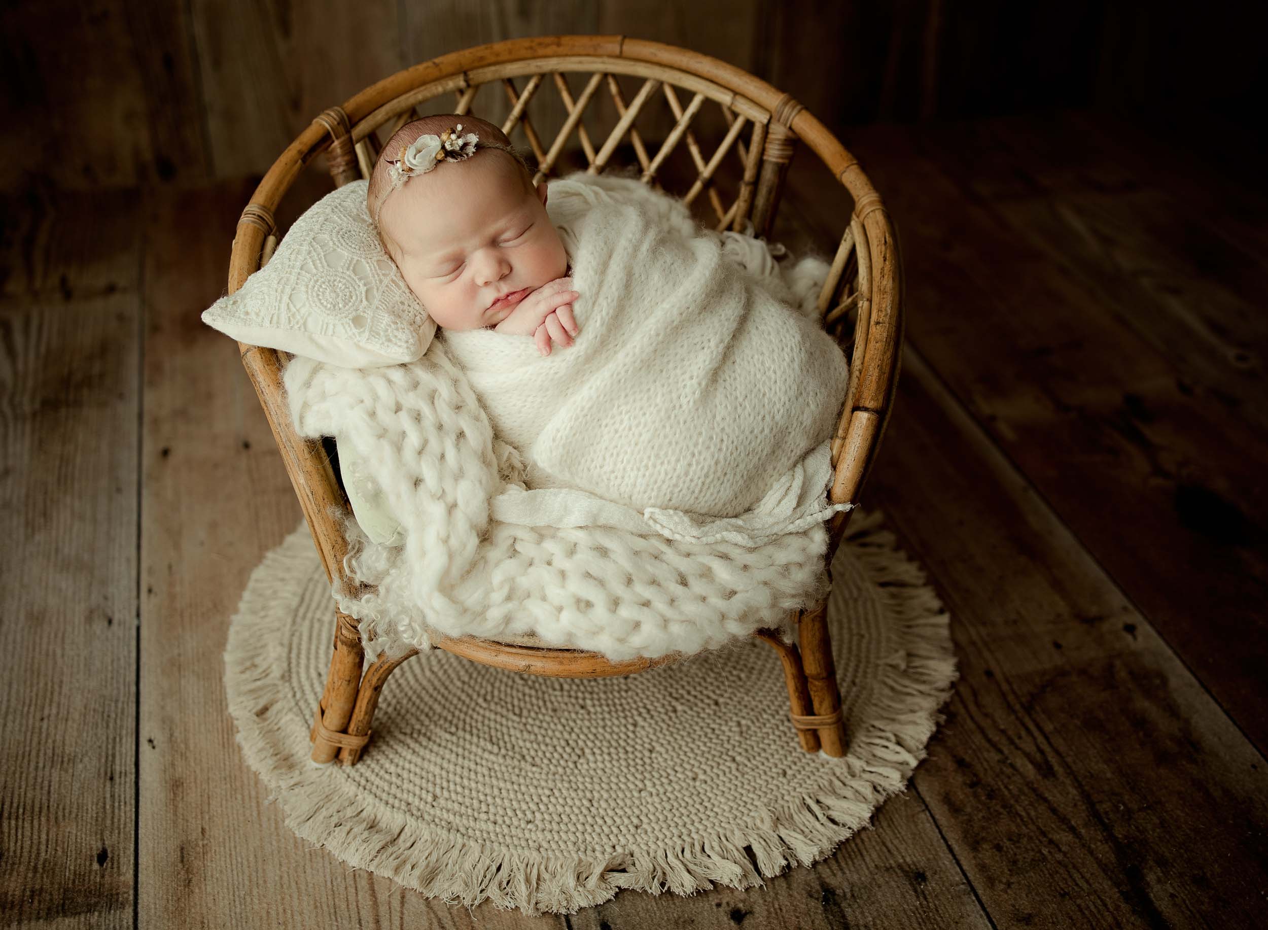Newborn baby girl wrapped in knit blanket propped up on a bamboo chair.