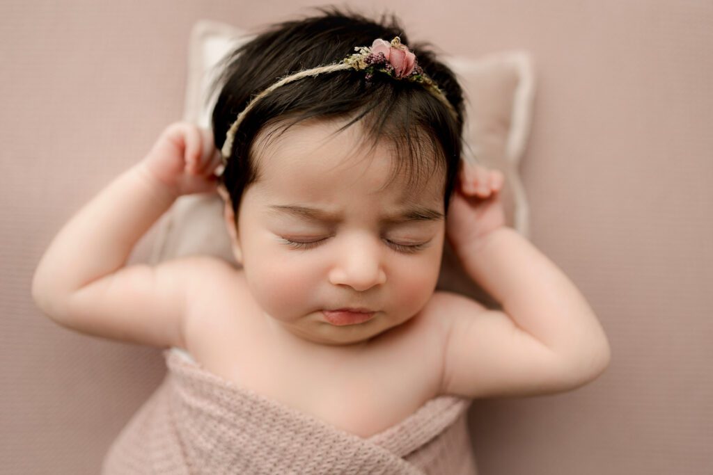 baby girl lying on soft pink backdrop with head on baby sized pillow during newborn photoshoot.