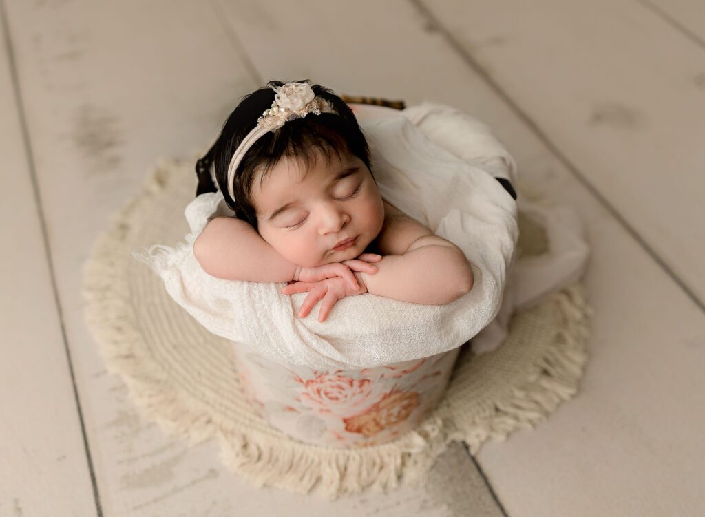 Baby girl with lots of dark hair posed in a bucket painted with pastel flowers on a cream floor. 