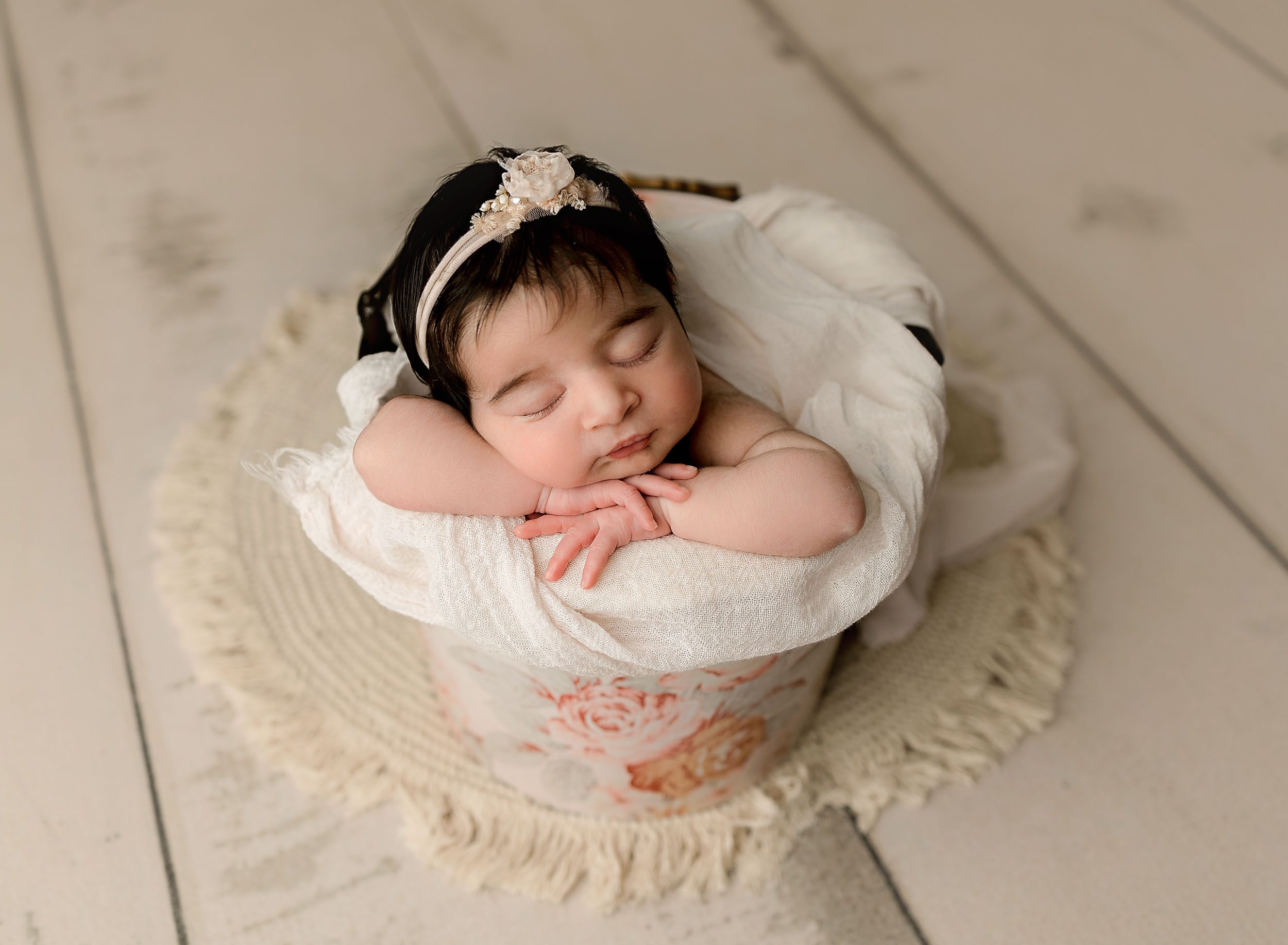 Baby girl with lots of dark hair posed in a bucket painted with pastel flowers on a cream floor.