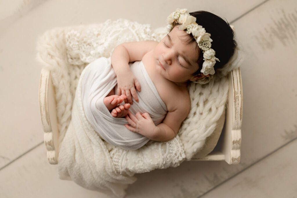 Baby girl with floral headband half wrapped in stretchy cream fabric lying in a cream wooden baby crib. 