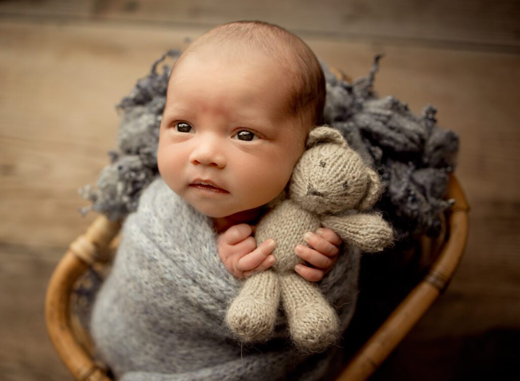 awake newborn baby boy swaddled in gray blue wrap holding a teddy bear in a bamboo basket prop.