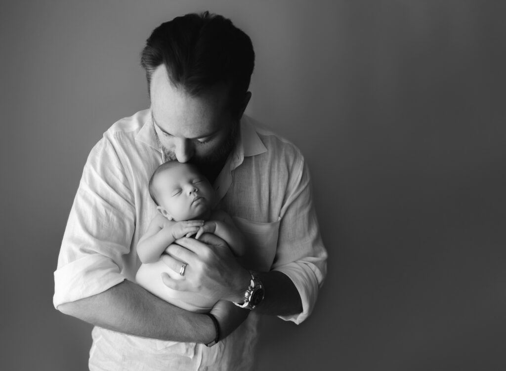 black and white photo of dad holding his newborn baby boy in front of his chest and kissing his head