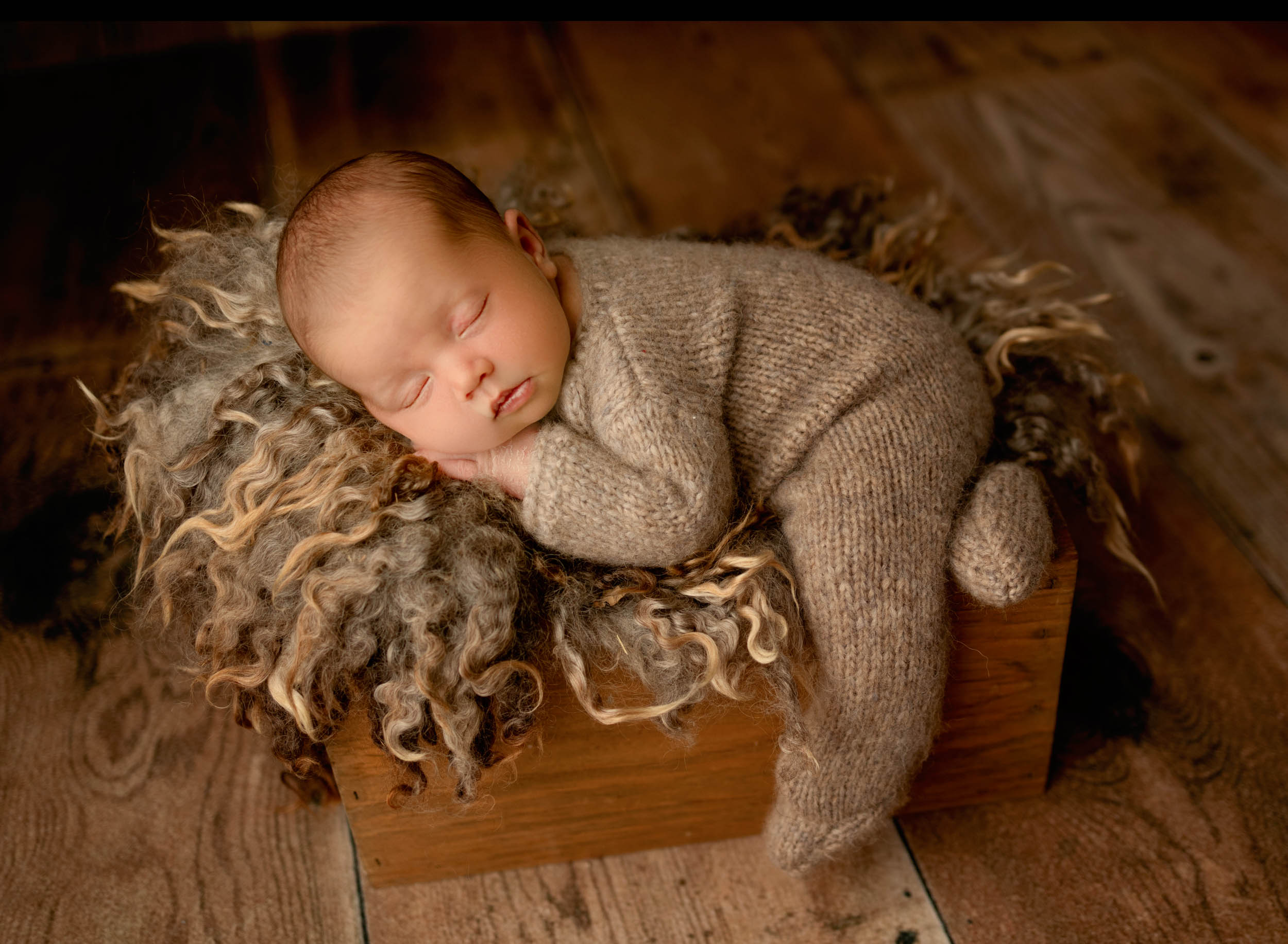 sleeping newborn baby boy wearing gray knit outfit laying in wood bed prop with fur blanket