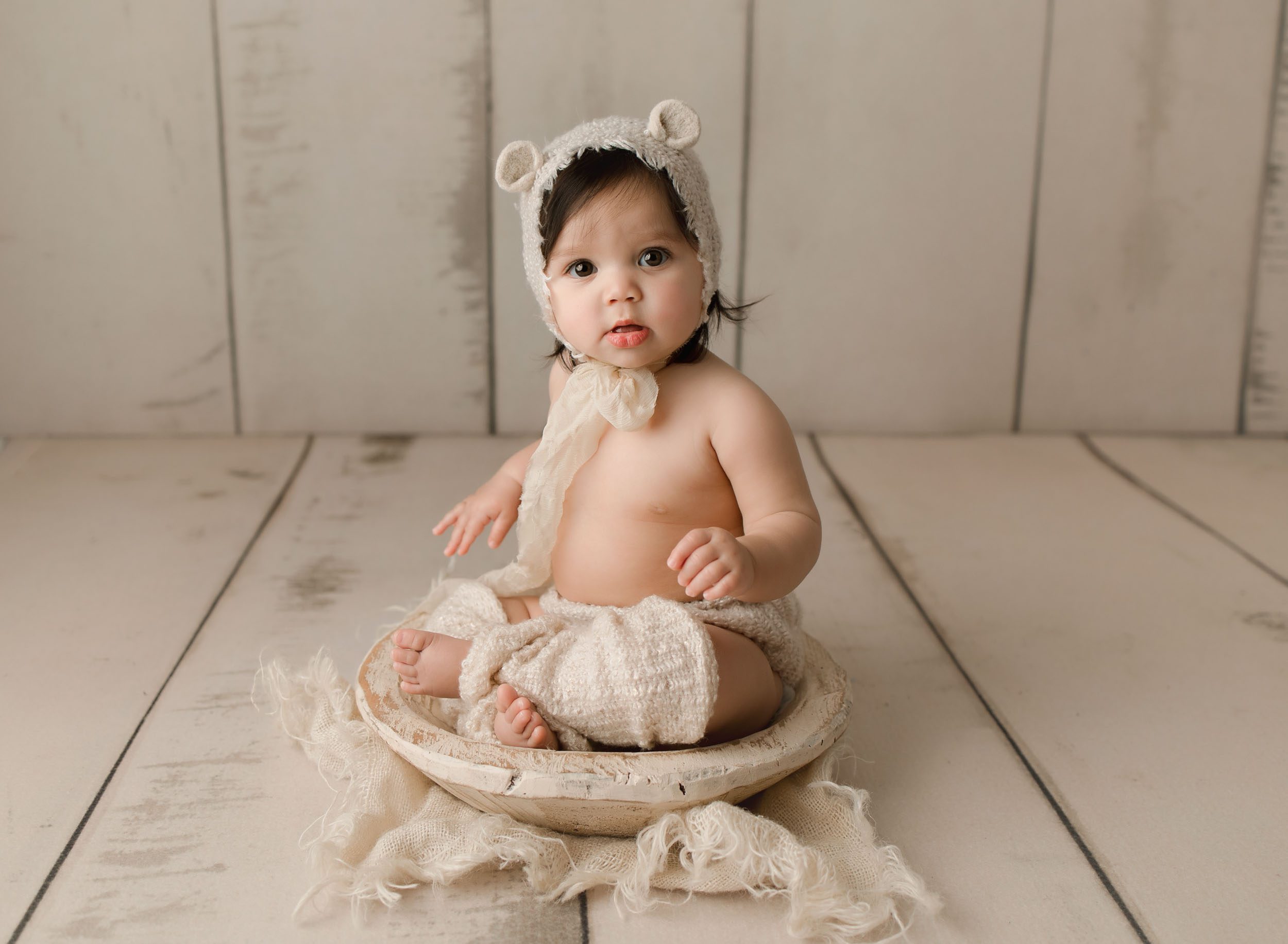 6 month old baby girl wearing teddy bear bonnet and leg warmers sitting in shallow cream colored bowl