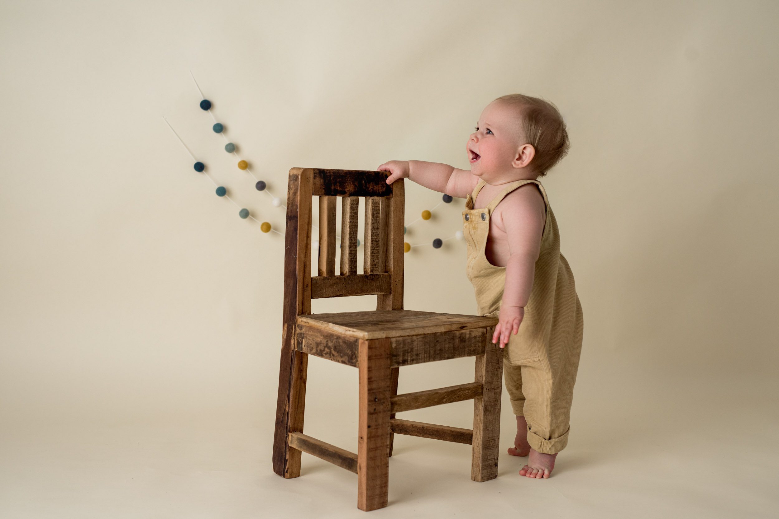 one year baby boy standing next to a chunky wooden chair