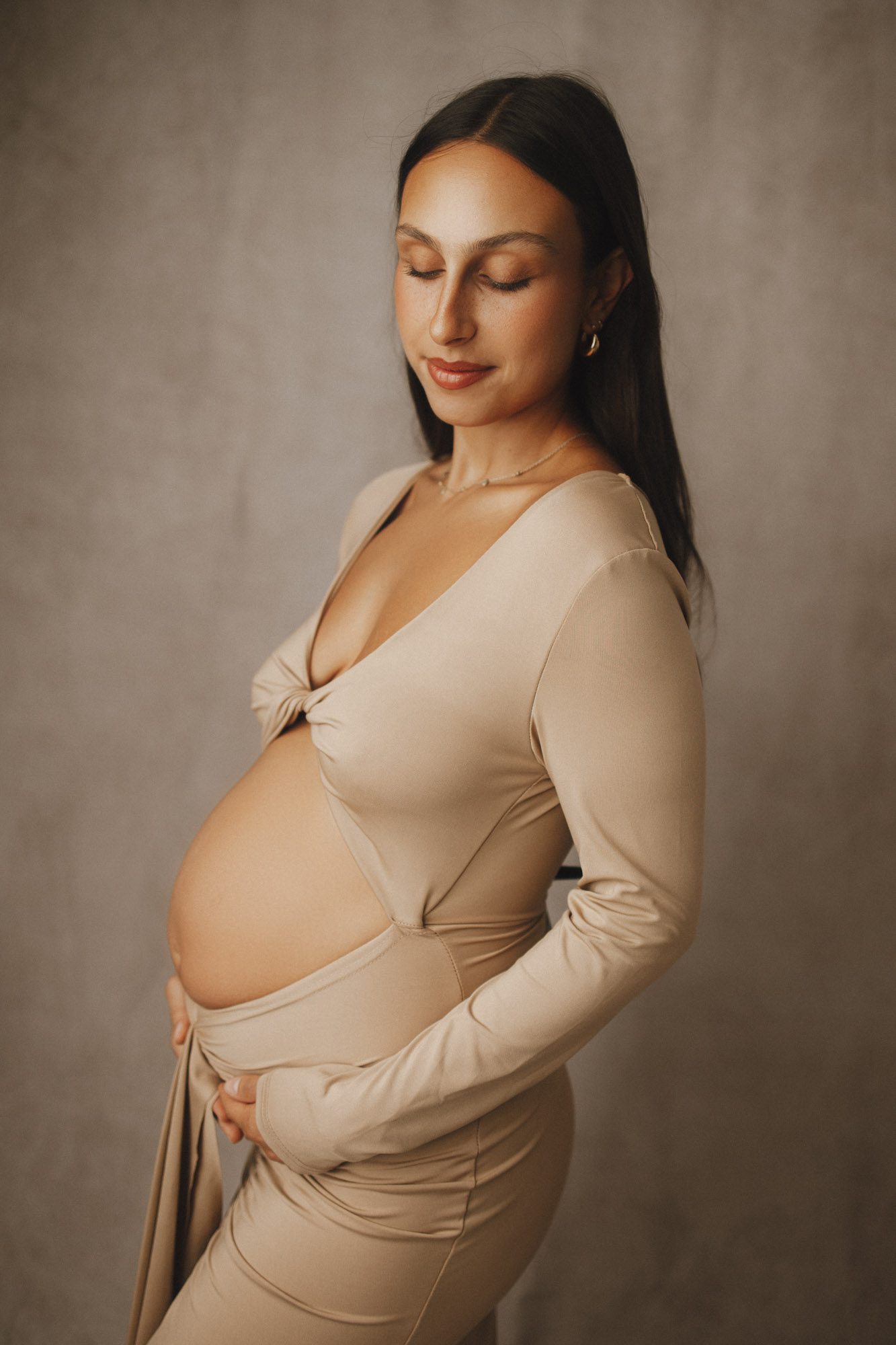 Side profile image of pregnant mom with beige backdrop and dress with her belly exposed