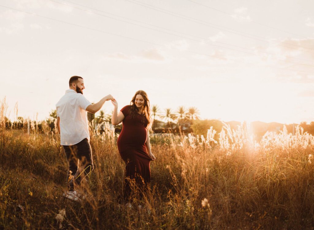 expectant parents with bold golden hour light walk through tall grass