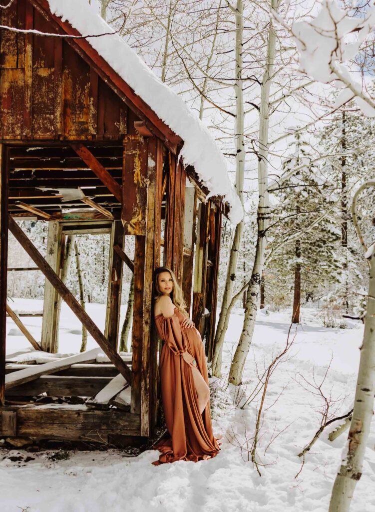 pregnant woman leaning against a snow covered barn showing a winter maternity photoshoot idea