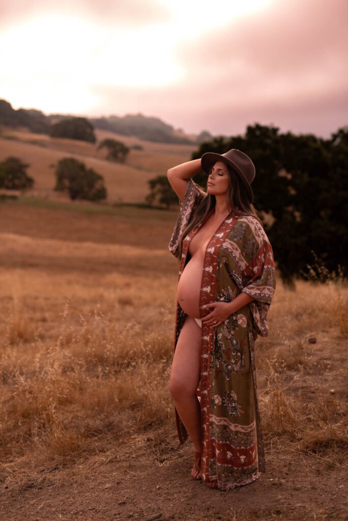 Expectant mom in golden field wearing open robe exposing baby bump wearing hat