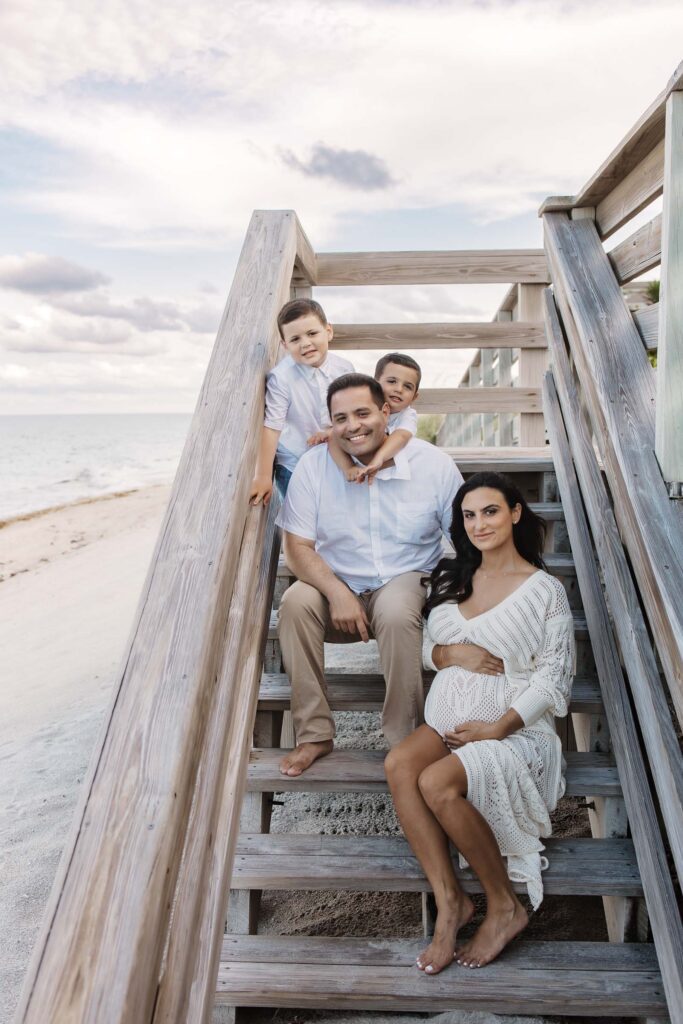 family with expecting mom sitting on the stairs at the beach