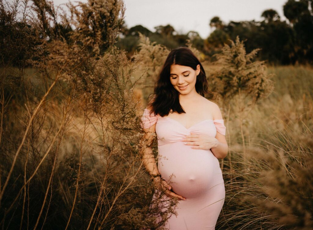 pregnant mom posing in field of tall golden grass looking at baby bump