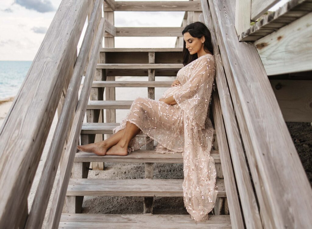pregnant woman in sparkly gown sitting on beach stairs
