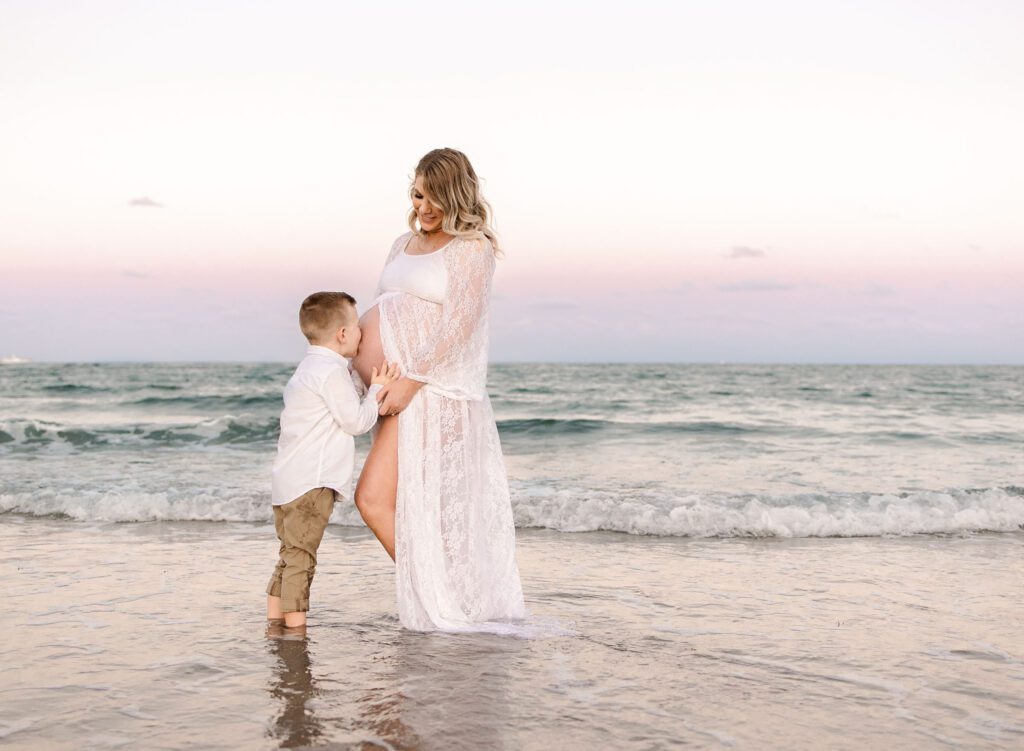 older sibling kissing mom's baby bump on the beach showing pregnancy photo ideas