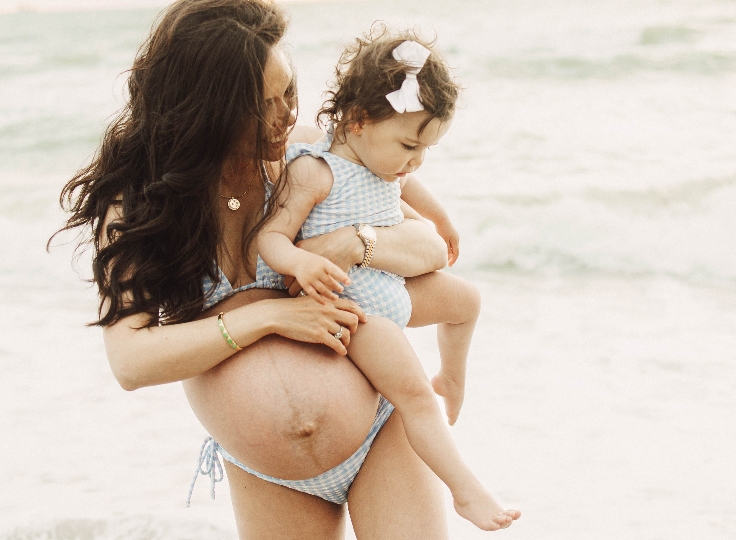 Toddler being held by her mom both wearing matching bathing suits. Mom is expecting and they are at the beach
