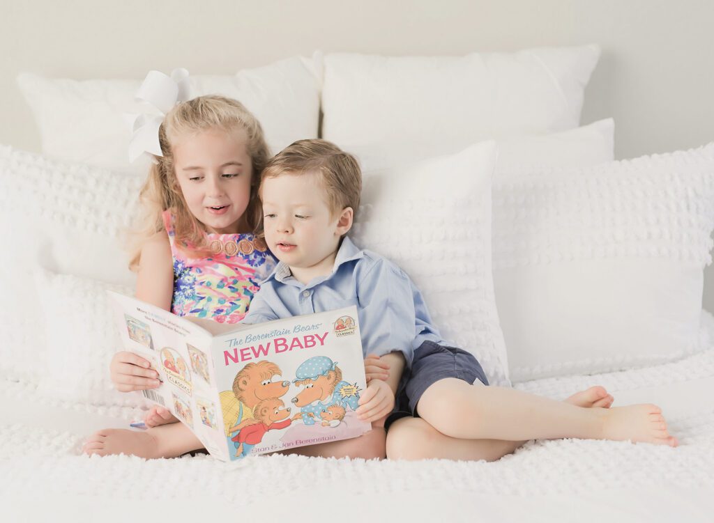 siblings reading new baby book on bed with white pillow during pregnancy announcement photos shoot