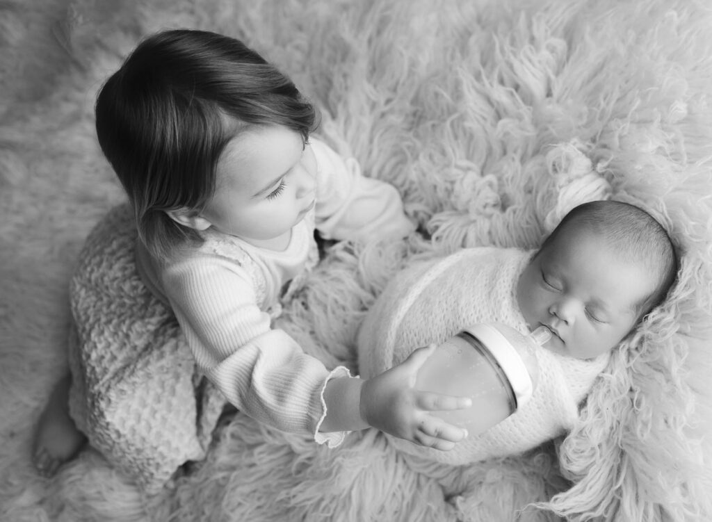 2 year old sister feeding bottle to newborn baby boy lying on flokati rug at his Coral Springs newborn photoshoot