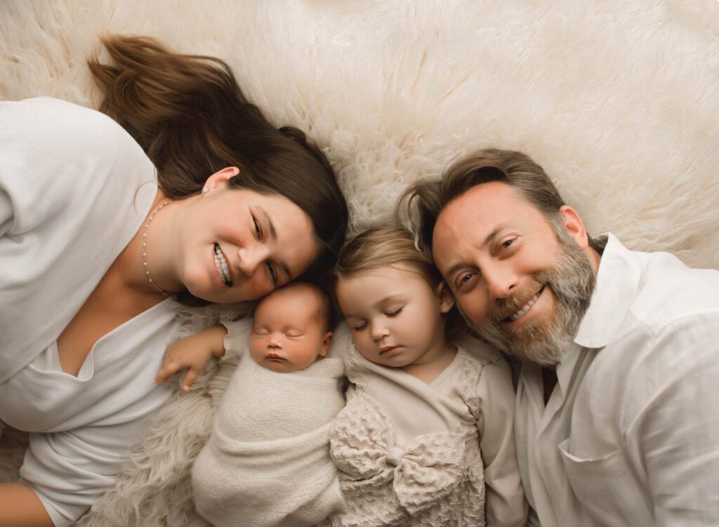 family of four laying down on cream fabric looking up at the camera with two sleeping babies between parents