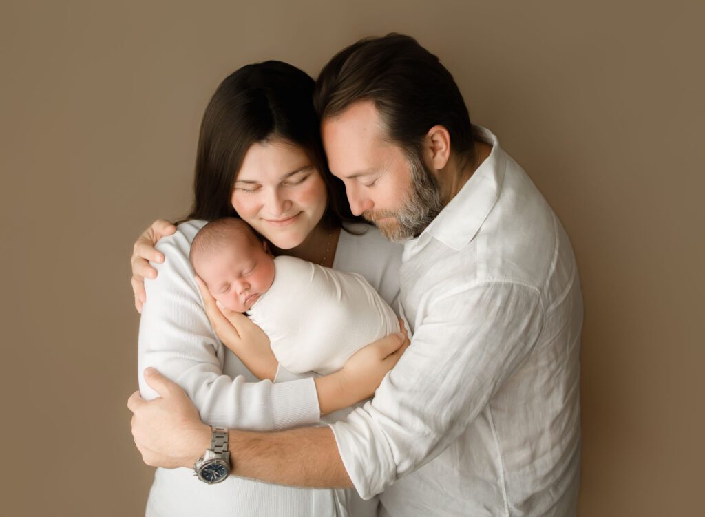 dad embracing mom holding their newborn baby looking down at him lovingly at their coral springs newborn photoshoot.